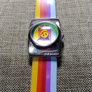 May include: A vintage JOE BOXER watch with a silver-tone square face and a rainbow-striped band. The watch face features a smiley face design. The watch band is a vibrant array of colors, including yellow, orange, red, purple, and blue.