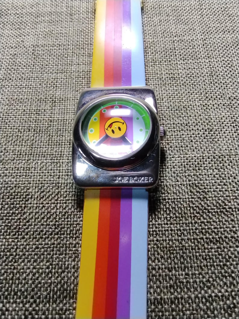 May include: A vintage JOE BOXER watch with a silver-tone square face and a rainbow-striped band. The watch face features a smiley face design. The watch band is a vibrant array of colors, including yellow, orange, red, purple, and blue.