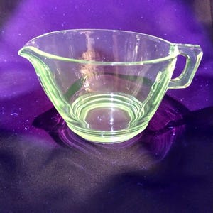May include: A clear, light green glass creamer with a handle and a pouring spout. The creamer has a rounded bowl shape and sits on a flat base. The glass has a slight glow, and the background is a dark purple.