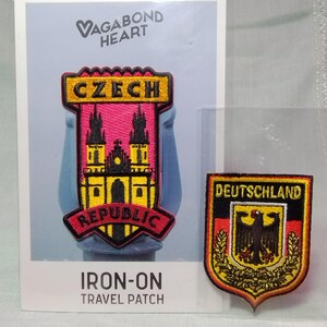 May include: Two embroidered iron-on travel patches. One rectangular patch features the text CZECH REPUBLIC with a yellow and black church illustration on a pink background. The other shield-shaped patch displays DEUTSCHLAND with a black eagle.