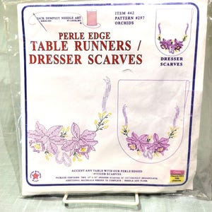 May include: A package of Jack Dempsey Needle Art table runners and dresser scarves. The package features a design of embroidered orchids in purple and yellow. The text reads "Perle Edge Table Runners / Dresser Scarves."