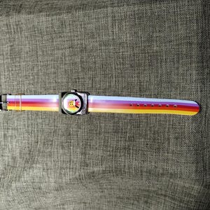 May include: A colorful wristwatch with a rainbow-striped band. The watch face is square with a cartoon character. The band has a gradient of colors including yellow, orange, red, purple, and white. The watch is set against a gray textured background.