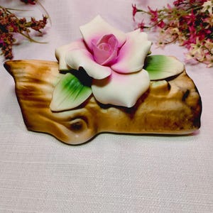 May include: A ceramic decorative figurine featuring a realistic rose with white and pink petals and green leaves resting on a brown, textured log-shaped base. The rose is centered on the log, which has a smooth, glossy finish and natural wood-like details.