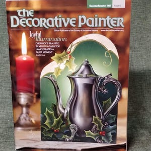 May include: The November/December 2002 issue of The Decorative Painter magazine features a cover illustration of a silver teapot surrounded by green holly leaves and red berries, with a red candle in a silver holder to the left of the teapot.