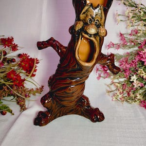 May include: A glossy brown ceramic vase shaped like a tree trunk with a wide, open-mouthed face. The figure has textured bark-like details, a hollow top opening, and outstretched arms. The interior of the mouth and the top opening are tan.