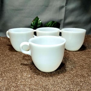 May include: Four white ceramic coffee cups with handles are arranged on a brown surface. The cups are evenly spaced, with one in the foreground and three behind it. The cups have a simple, rounded design.