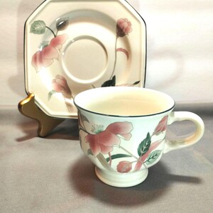 May include: A cream-colored teacup and saucer set. The cup and saucer feature a floral design with pink flowers, green leaves, and black trim. The saucer is octagonal. The cup has a pedestal base and a curved handle.