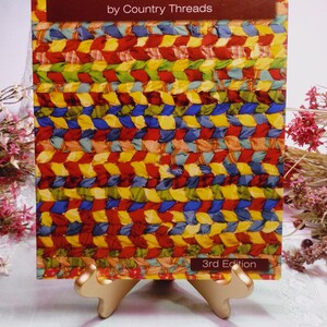 May include: A softcover book titled Rugs From Rags by Country Threads, 3rd Edition. The cover features a vibrant, multi-colored braided fabric pattern in shades of red, yellow, blue, and green. The book is displayed on a gold-toned metal stand.