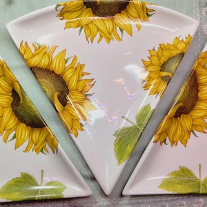 May include: Three triangular ceramic plates featuring a white background with a vibrant sunflower pattern. Each plate displays a large yellow sunflower with a dark brown center and green leaves, arranged in a repeating floral design across the surface.