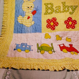May include: A quilted baby blanket featuring a yellow gingham border with ruffles. The design includes a yellow gingham bunny holding a carrot on a blue background, the text Our Baby in red and green, red flowers, and a train with three cars.