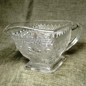 May include: A clear, square-shaped glass creamer with a handle and spout. The creamer features a detailed, embossed pattern of diamonds, flowers, and other shapes. The creamer sits on a square base and is likely vintage.