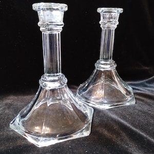 May include: Two clear glass candle holders. Each has a square base that tapers up to a cylindrical stem, and a flared top. The glass is transparent, allowing light to pass through. The candle holders are likely for decorative use.