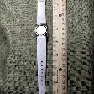 May include: A rectangular silver-toned watch with a white band featuring the text "STR CASE" in pink and green. The watch is next to a wooden ruler marked in inches.