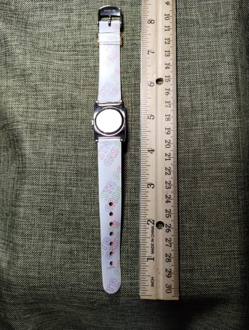 May include: A rectangular silver-toned watch with a white band featuring the text "STR CASE" in pink and green. The watch is next to a wooden ruler marked in inches.