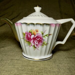 May include: A white porcelain teapot featuring a ribbed body, a gold-trimmed spout, handle, and lid finial. The upper rim is decorated with a pink and gold scalloped pattern, while the center displays a floral motif of pink roses and green leaves.