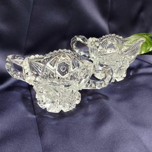 May include: Two clear, cut-glass serving dishes. One is a sugar bowl with handles and a lid, and the other is a creamer with a spout and handle. Both have intricate geometric patterns and sit on ornate bases. The background is a dark blue fabric.