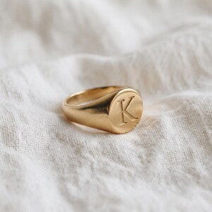 Può includere: Un anello a sigillo color oro caratterizzato da una superficie liscia e lucida con la lettera maiuscola K incisa sulla parte circolare. L'anello presenta una fascia solida e arrotondata che si restringe leggermente verso il retro.