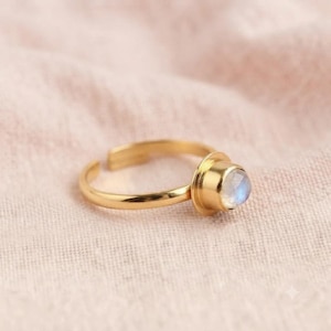 Può includere: Un anello color oro con una pietra cabochon rotonda, liscia e iridescente incastonata in una lunetta. La fascia è aperta sul retro, consentendo la regolazione, e il metallo ha una finitura lucida che riflette la luce sulla sua superficie.