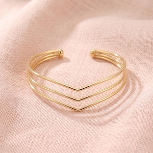 Puede incluir: Una pulsera tipo brazalete de metal dorado con tres bandas paralelas que convergen en una forma de V central. El diseño abierto termina en dos pequeñas esferas redondeadas en cada extremo, creando una estructura geométrica.