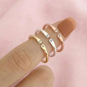 Op de afbeelding: Vier verstelbare ringen in goud, zilver en roségoud, elk met een gegraveerde initiaal. De ringen worden op een vinger getoond tegen een zachtroze achtergrond. De initialen zijn "S" en "H".