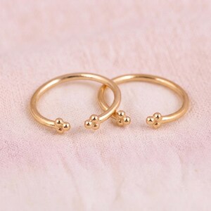 May include: Two gold-toned open rings, each featuring a small decorative cluster of three tiny spheres at both ends of the band. The rings have a smooth, polished finish and a circular shape with a gap between the ends, creating a delicate appearance.