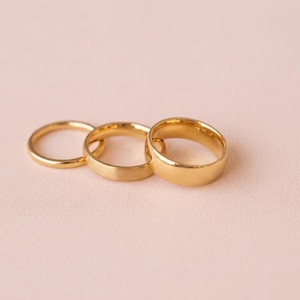Puede incluir: Tres anillos de oro de diferentes anchos están dispuestos sobre una superficie rosa claro. Los anillos tienen un acabado pulido, uno de ellos con una textura mate. Los anillos son probablemente para compromiso o boda.