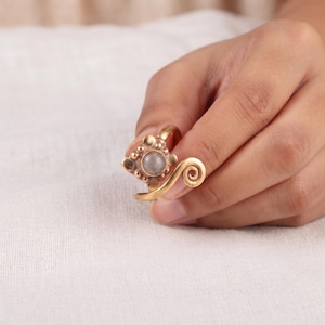 Op de afbeelding: Een goudkleurige metalen ring met een centrale ronde grijze steen in een vierkant montuur versierd met kleine bolvormige accenten. De ringband is open en eindigt in een decoratief spiraalontwerp aan één kant van de zetting.