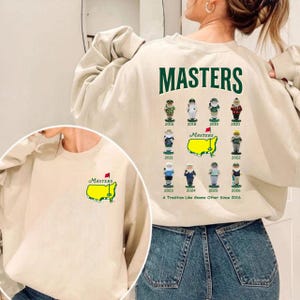 Peut inclure: Sweat-shirt crème avec le mot "MASTERS" en lettres vertes. Le dos présente des illustrations de gnomes avec les années de 2016 à 2026. Une petite image du logo Masters est présente sur le devant. Le texte "A Tradition Like Gnome Other Since 2016" est également présent.