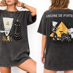 May include: Dark gray t-shirt featuring a graphic of two stylized masks with white polka dots and gold accents. The back of the shirt displays the text "ANGINE DE POITRINE" above a band illustration with a yellow triangle.