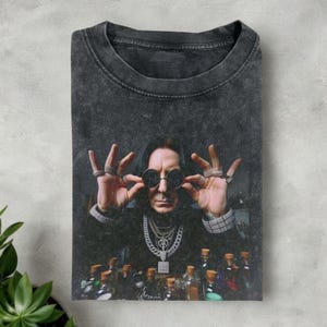 May include: Dark gray t-shirt featuring a portrait of a man wearing round sunglasses and multiple rings. The man is wearing a black outfit with multiple necklaces. Below the man are several bottles with brown liquid.
