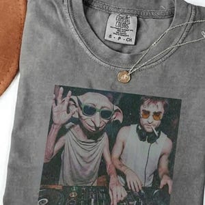 May include: Gray Comfort Colors t-shirt featuring a graphic of two figures with sunglasses at a DJ setup. A silver necklace with a pendant is on the shirt. The shirt has a tag that says "Comfort Colors".
