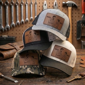 May include: Several trucker caps in a stack, with colours including grey, black, white, and camouflage. Each cap has a leather patch with the text "DADDY MAX" and a father-son illustration. The hats are displayed on a wooden surface.