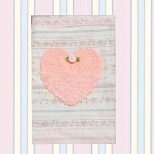 May include: A rectangular fabric piece featuring a white background with horizontal stripes of pink and blue floral patterns. A large peach-colored heart with white polka dots and a pink fabric rose sits in the center, partially covered by lace.