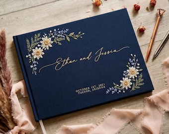 Puede incluir: Un libro de visitas cubierto de tela azul marino con flores de margarita blancas y amarillas bordadas y follaje verde. La cubierta muestra el texto "Ethan and Jessia" en letra cursiva dorada, seguido de "OCTOBER 1ST, 2021" y "TUCSON, ARIZONA" en oro.