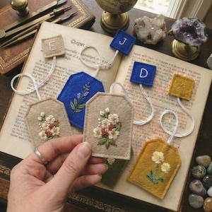 May include: A collection of handmade felt bookmarks with embroidered floral designs and lettered tags. The bookmarks are in various colors, including beige, blue, and yellow, and are attached to the tags with white string.