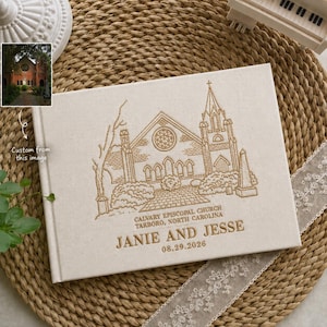 May include: A rectangular off-white fabric-covered guest book featuring a gold-embroidered line drawing of Calvary Episcopal Church in Tarboro, North Carolina, with the names Janie and Jesse and the date 29.08.2026 stitched below the building.