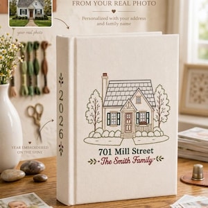 May include: A cream-colored fabric photo album featuring an embroidered illustration of a house on the front cover. The text 701 Mill Street and The Smith Family is embroidered below the house. The year 2026 is embroidered on the album spine.