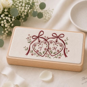 May include: A rectangular wooden box with a cream-colored fabric lid featuring embroidered red ribbons, green vines, and small pink flowers. Two heart-shaped ornaments are stitched in the center, each containing the embroidered letters T and H.
