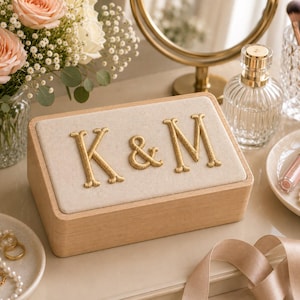 May include: A rectangular wooden jewelry box with a light beige fabric lid featuring the embroidered gold letters K & M. The box has a smooth, natural wood finish and a clean, simple design, serving as a functional container for small personal items.
