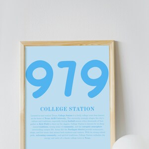 May include: A framed poster with a light blue background features the number "979" in large, bold, blue font. Below, the text "COLLEGE STATION" is printed in a smaller, darker blue font. The poster is in a light wood frame.