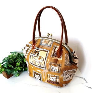 以下が含まれることがあります： 猫柄の茶色のハンドバッグ。キスロック開閉と茶色のハンドルが特徴です。バッグの生地には、さまざまなポーズの猫のイラストがフレームに入って表示されています。ハンドルは光沢のある茶色で、バッグはヴィンテージ感があります。