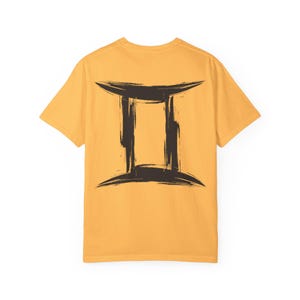 Peut inclure: T-shirt jaune moutarde avec un symbole du zodiaque Gémeaux marron foncé au dos. Le symbole est un design au trait de pinceau. Le t-shirt a des manches courtes et un col rond.