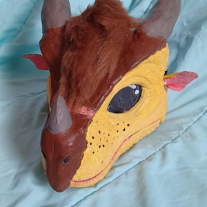 May include: A handmade dragon mask with a yellow face, brown horns, and a brown fur mane. The mask has a detailed design with black eye and red accents. The mask is designed to be worn.