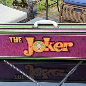 May include: A rectangular wooden box with a purple front panel featuring the words "THE Joker" in yellow and orange lettering. The box has a green and red trim, a white handle, and red latches. The Joker's face is in the center of the word "Joker".