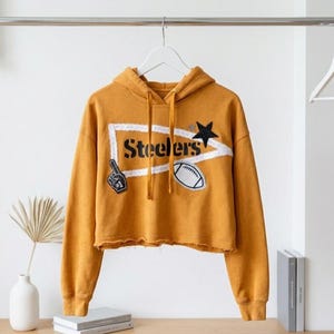 May include: Mustard yellow cropped hoodie with the word "Steelers" in black letters inside a white outlined shape. The hoodie has a black star and football patch, and a foam finger patch. The hoodie is hanging on a white hanger.