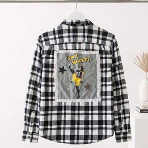 Puede incluir: Camisa de franela a cuadros blancos y negros con un parche decorativo que presenta a un jugador de fútbol americano en amarillo y negro. El parche incluye el texto "SAY WATT!" y estrellas. La camisa está colgada en una percha blanca.