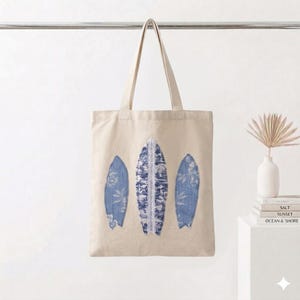 May include: A beige canvas tote bag featuring three blue surfboard designs. The surfboards have floral and patterned details, with a white lace accent on the center board. The bag has two handles and is hanging against a white background.