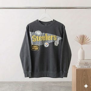 May include: A charcoal gray sweatshirt with the word "Steelers" in gold lettering on a pennant-shaped patch. Additional patches include a football, a logo, and the phrase "Pittsburgh is for winners."