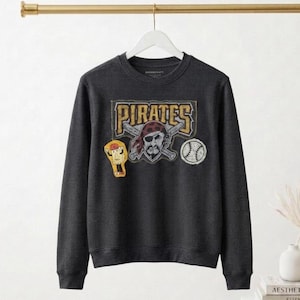 May include: Dark gray crew neck sweatshirt with a textured waffle knit. The front features a distressed graphic of the word "PIRATES" above a pirate with crossed bats and a baseball. A cartoon character and baseball patch are also visible.