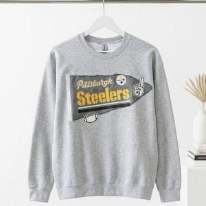 May include: A gray sweatshirt featuring the Pittsburgh Steelers logo. The logo includes the team name in yellow and black, a football, and a pointing finger with the number one. The sweatshirt is hanging on a white hanger.